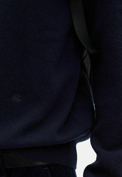 Marineblauer Pullover mit weicher Textur, mit einem kleinen Logo auf der Brust gestickt, kombiniert mit dunklen Hosen und einem schwarzen Gürtel.