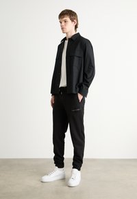 Zwart overhemd met knoopsluiting en twee borstzakken, gecombineerd met zwarte sweatpants met een logo en witte sneakers met een dikke zool.