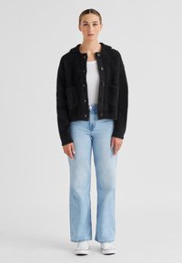 Schwarze, flauschige Strickjacke mit Kragen, Knopfleiste, zwei vorderen Taschen; kombiniert mit hellblauen Schlagjeans und weißen Turnschuhen.