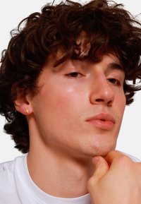 Orecchino a forma di croce in oro su un modello maschile. Il modello ha capelli ricci scuri e una pelle con texture naturale, indossa una maglietta bianca. Inquadratura ravvicinata.