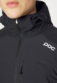 Giacca nera impermeabile con colletto alto, chiusura con zip sul davanti e tasca laterale. Presenta un logo sulla spalla e un tessuto testurizzato.