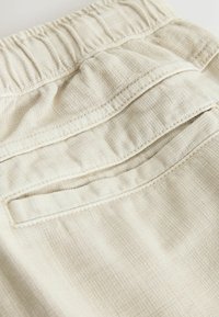 Gros plan sur un pantalon en tissu beige texturé avec une taille élastique et une poche arrière passepoilée avec coutures visibles.