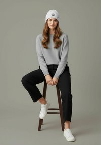 Gray knit sweater, black tapered trousers, and white sneakers. The model sits on a wooden stool with a light gray background.