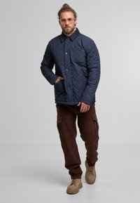 Gepolsterte marineblaue Jacke mit Kragen, Vordertasche und Druckknopfverschluss. Kombiniert mit braunen Corduroy-Cargohosen und tanfarbenen Schnürstiefeln.