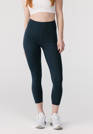 Kvinne iført svarte korte leggings, hvit cropped topp og hvite treningssko, stående foran en ensfarget lys bakgrunn.