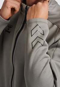 Chaqueta atlética gris con cremallera frontal. Presenta un tejido texturizado y un diseño de chevrón negro en la manga derecha. Logo visible en la parte delantera.
