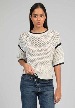 Jeune femme aux longs cheveux foncés portant un pull en maille blanc à manches courtes avec bordure noire et un jean bleu, debout devant un fond neutre.