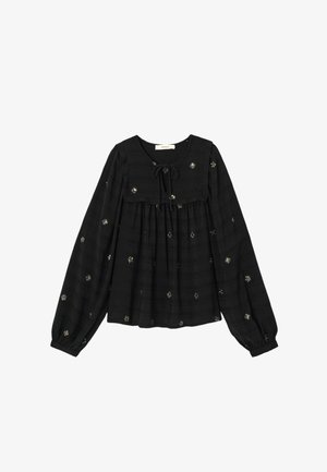 Blouse noire à manches longues avec un col rond et un détail de nœud, ornée de motifs d'étoiles brodés en argent sur un tissu texturé.