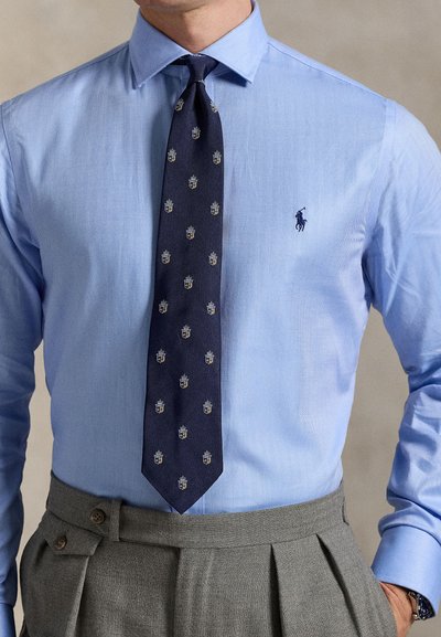 Cămașă albastru deschis, cu model espiga, și cravată bleumarin cu model repetitiv de scuturi. Pantaloni gri, plisati, cu nasturi pe lateral.