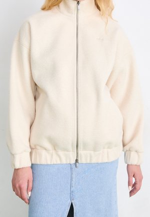 Femme portant une veste polaire crème à fermeture éclair avec poignets et ourlet élastiqués, associée à une jupe en denim bleu clair avec une fente à l'avant.