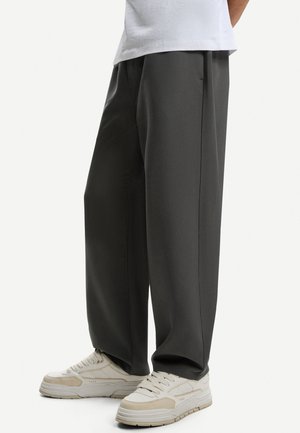 Person iført løstsiddende mørkegrå bukser og hvide sneakers, stående mod en ensfarvet lys baggrund.
