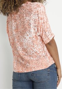 Blusa leggera a maniche corte con un motivo maculato corallo e pesco e dettagli con bottoni sulle maniche, indossata con jeans in denim blu.