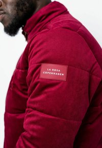 Giacca in velluto a coste rossa con una texture trapuntata e una toppa con il logo "LA ROSA COPENHAGEN" sul braccio sinistro. Design con colletto alto.