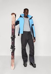 Smučarska oprava z modro puhovko, sivim puloverjem, črnimi hlačami, ki drži rjave smuči s črnimi vezmi in črne smučarske čevlje.