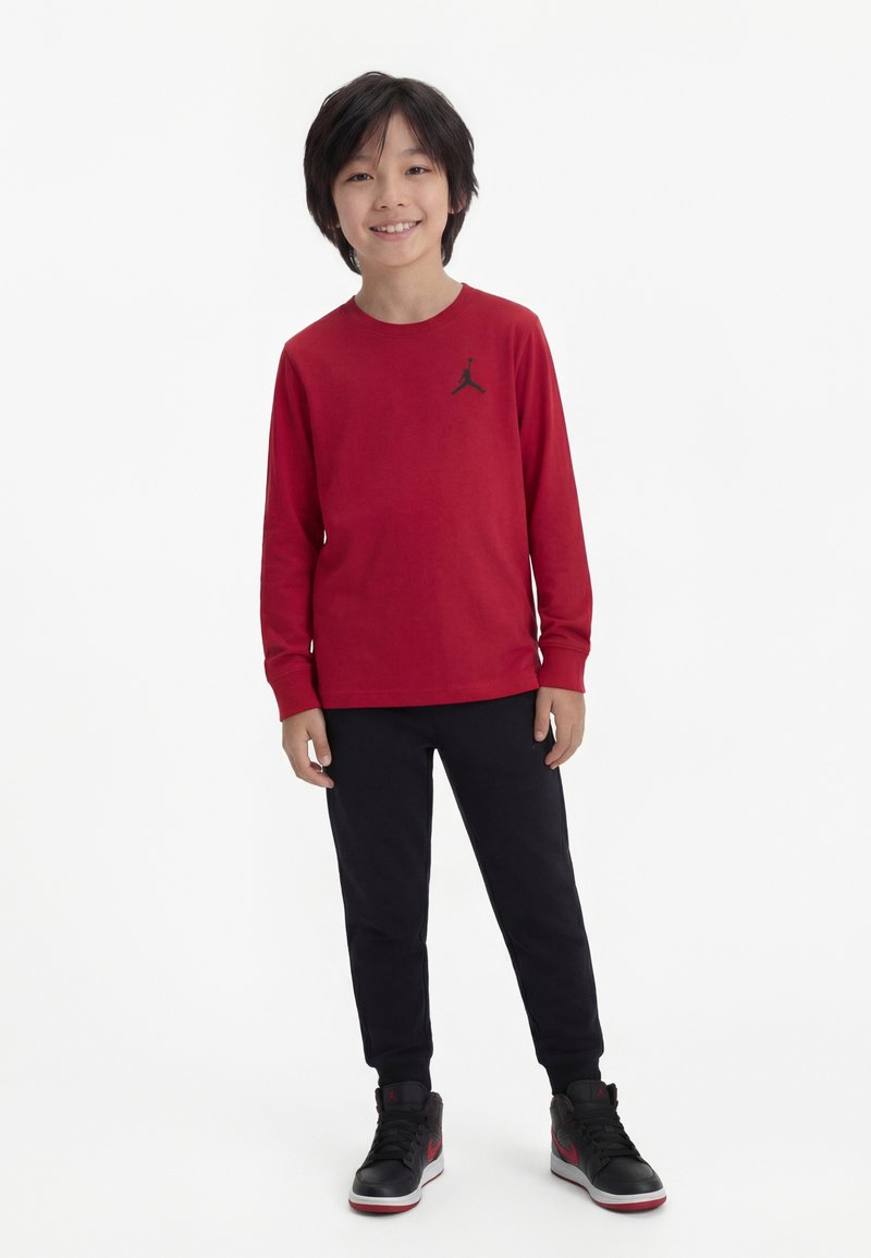 Băiat stând zâmbind, purtând o bluză roșie cu mâneci lungi și logo negru, pantaloni negri și adidași negri cu accente roșii pe un fundal alb.