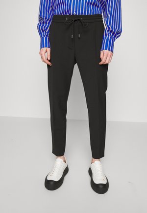 Homme portant un pantalon noir à cordon, des baskets blanches avec semelles noires et une chemise à manches longues bleue à rayures, debout devant un fond uni.