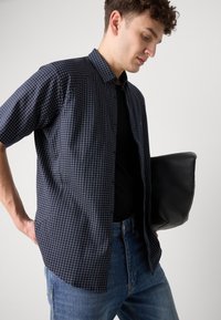 Αμάνικο πουκάμισο με ναυτική καρό, φτιαγμένο από βαμβάκι, συνδυασμένο με μαύρη μπλούζα και μπλε τζιν, κρατώντας μια μαύρη δερμάτινη τσάντα κλάσης.