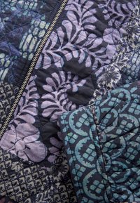 Close-up van een gewatteerde jas met een koperen rits, met blauwe, paarse en zwarte bloemen- en geometrische patronen.