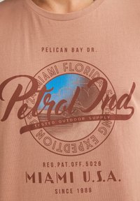 Hnědé tričko s kulatou grafikou, která obsahuje modrý a hnědý text: "Petrol Ind," "Miami Florida," a "Tested Outdoor Supply." Měkký materiál.