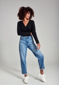 Sort v-hals cardigan, blå højtaljede jeans med opsmøg, og hvide sneakers. Krøllet hår stylet løst, stående positur.