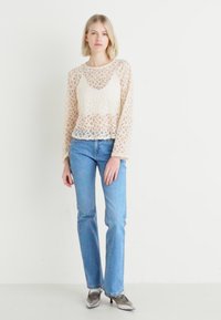 Top de encaje color crema con mangas largas, escote redondo, patrón de círculos en capas, combinado con jeans azul claro y zapatos de tacón metálicos.