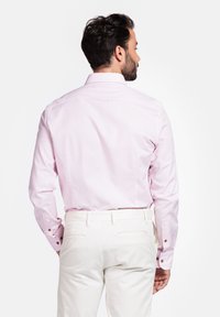 Lichtroze overhemd met lange mouwen en een button-down kraag. Heeft knoopaccenten op de manchetten. Draagt met witte broek, met een nette achterkant te zien.