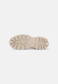 Semelle d'une chaussure en beige clair, présentant un design texturé et robuste avec des crampons distincts pour l'adhérence et une zone centrale lisse.