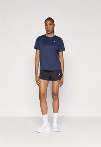 Ναυτική αθλητική μπλούζα φτιαγμένη από ελαφρύ ύφασμα, συνδυασμένη με μαύρες κοντές βερμούδες. Το μοντέλο φοράει λευκά παπούτσια και κάλτσες μέχρι τον αστράγαλο, στέκεται σε απλό φόντο.