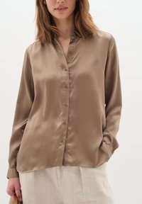 Camicia button-up in satin marrone con colletto e maniche lunghe, caratterizzata da una texture liscia e una leggera lucentezza, abbinata a pantaloni in lino di colore chiaro.