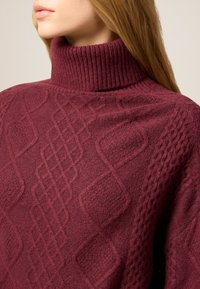 Vinröd polotröja i texturerad stickning med diamant- och flätmönster, med hög ribbad krage och mjukt tyg.