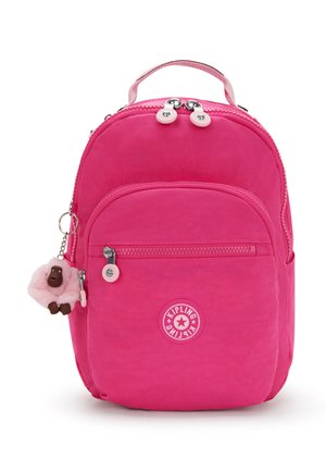 Jasnoróżowy plecak Kipling z przednią kieszenią na zamek, górnym uchwytem i małą pluszową przywieszką w kształcie małpki przymocowaną do bocznego zamka.