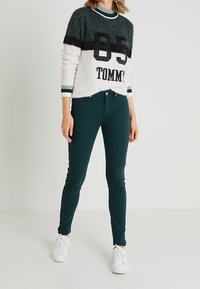 Suéter fuzzy blanco y verde con rayas negras y estampado "TOMMY", combinado con pantalones ajustados verde oscuro y zapatillas blancas.