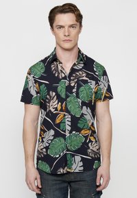 Camisa de manga corta hecha de tela con un fondo azul marino y grandes patrones de hojas en verde y marrón, y un bolsillo delantero con botones de madera.