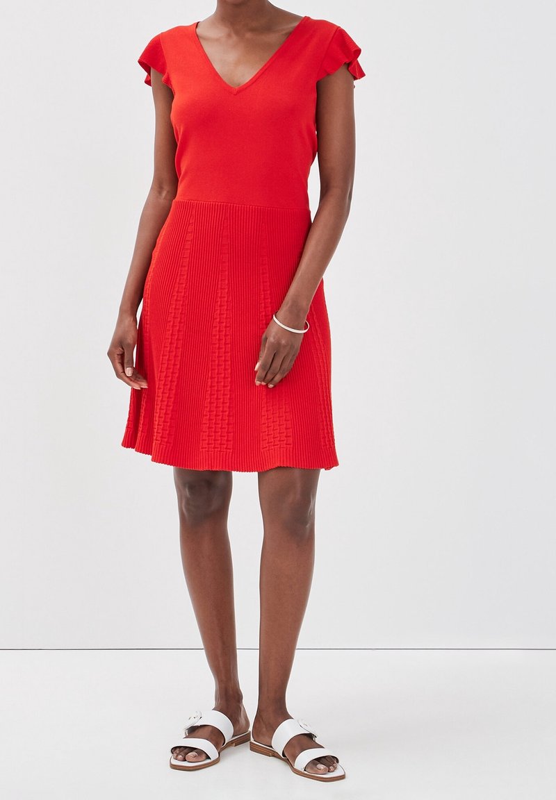 Robe tricotée rouge avec un décolleté en V et de courtes manches volantées, dotée d'une jupe plissée texturée. Assortie à des sandales blanches.