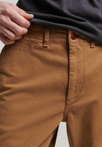 Pantalones de algodón marrón con corte recto, que cuentan con cierre de botón, bolsillos frontales y detalles de costura visibles. Textura suave.