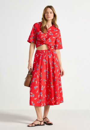 Nainen, jolla on punainen kukallinen lyhythihainen nyöritystoppiyläosa ja siihen sopiva midihame, pitää kädessään ruskeaa käsilaukkua ja hänellä on jalassa ruskeat sandaalit.