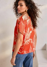 Red shirt with a relaxed fit, featuring a palm leaf pattern in cream. Short sleeves with cut-out accents, paired with blue jeans.