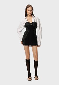 Schwarzes Mini-Kleid mit korsettartigem Oberteil und strukturiertem Stoff; kombiniert mit einem weißen Oversized-Hemd, durchsichtigen schwarzen Kniestrümpfen und Sandalen.
