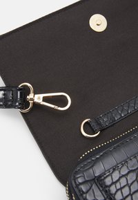 Černá texturovaná kožená taška uvnitř s zlatými kovovými doplňky, obsahující bezpečné zapínání na patentku, zip a místo pro odnímatelný popruh.