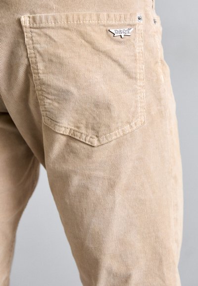 Un pantalon en velours côtelé beige montrant une poche arrière avec une épingle logo argentée DSQ2 sur la hanche droite, sur un fond clair.