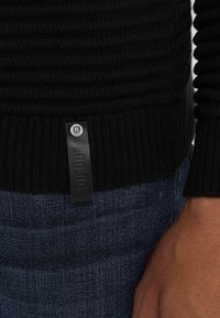 Zwarte geribbelde sweater met een leren label en metalen knop, gecombineerd met donkerblauwe denim jeans en een zichtbare hand aan de zijkant.