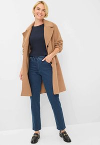 Manteau en mélange de laine marron, pull en maille côtelée bleu marine, jeans droits bleus et mocassins noirs avec accents en métal doré, sur fond blanc.