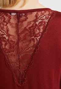 Burgundy topp med spetsbakgrund som har blommönster, en V-formad utskärning och en slät, stickad textur på huvuddelarna.