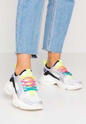 Białe masywne sneakersy z iryzującymi panelami i wielokolorowymi sznurówkami noszone z jasnoniebieskimi, krótkimi, postrzępionymi dżinsami na jasnym tle.