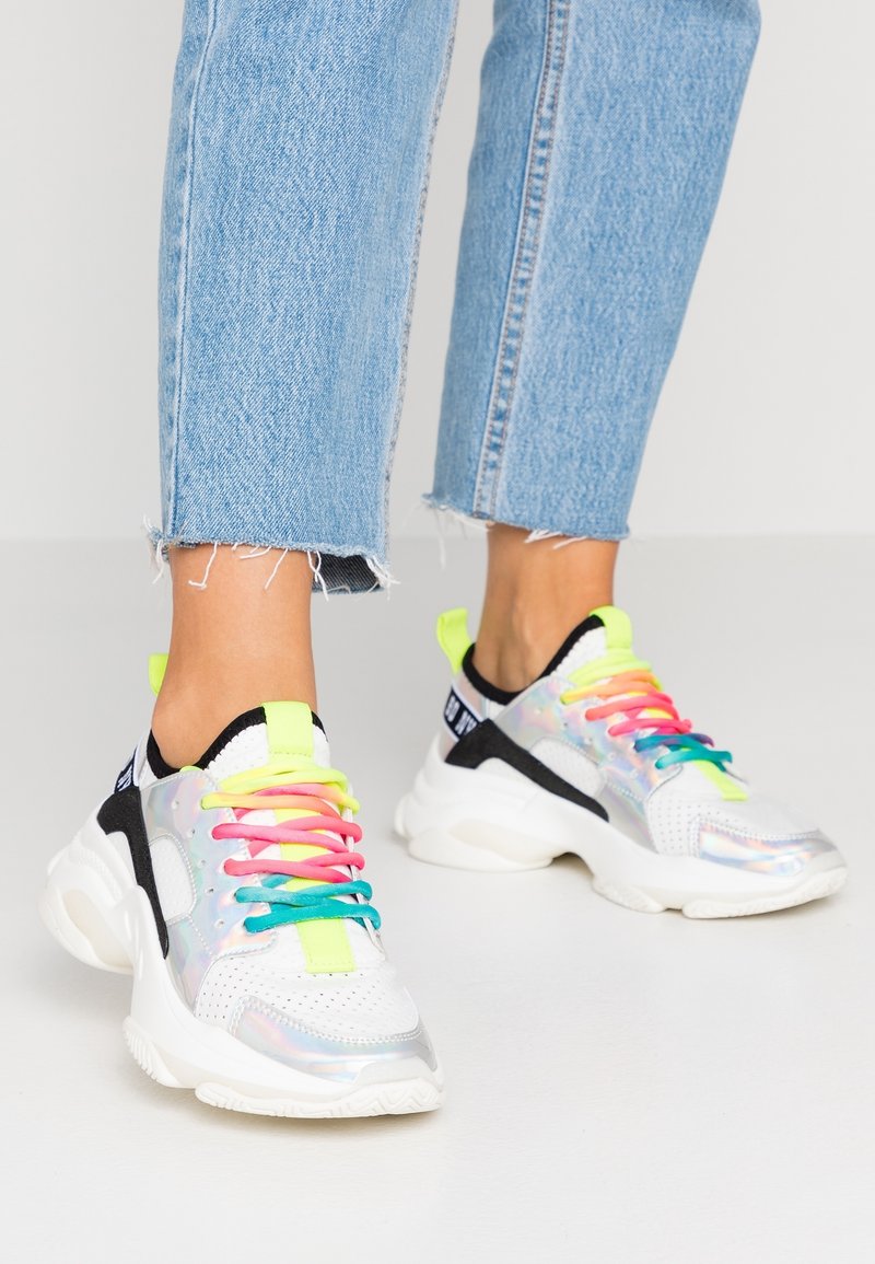 Baskets blanches épaisses avec des panneaux irisés et des lacets multicolores portées avec un jean court bleu clair effiloché sur un fond clair.