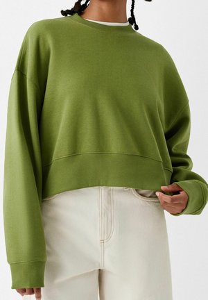 Person iført en oversized olivengrøn sweatshirt med ribkanter ved ærmer og bund, kombineret med højttaljede offwhite bukser.
