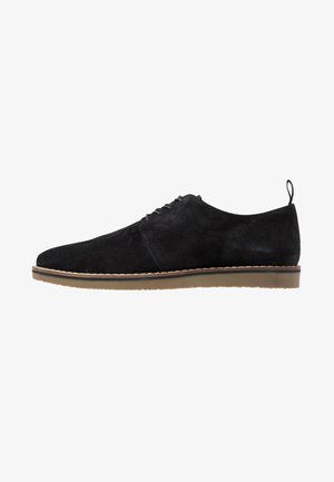 Chaussure à lacets pour homme en daim noir avec une semelle en caoutchouc beige et une languette à l'arrière, présentée en profil latéral sur un fond blanc.