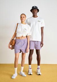 Weiße T-Shirts mit Logo, lavendelfarbene Shorts und Sportschuhe. Eine braune Schultertasche mit Fransen-Details. Neutraler Hintergrund und gelber Boden.