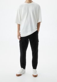 Witte oversized katoenen t-shirt, relaxte pasvorm, korte mouwen. Zwarte cargo broek met zijzakken, gestructureerde stof. Witte sneakers met bruine zolen.