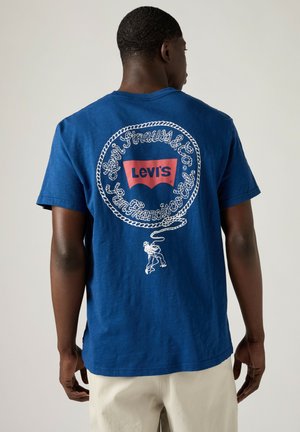 Hombre que lleva una camiseta azul Levi's con un círculo de cuerda blanca, logo rojo y gráfico de vaquero lanzando lazo en la espalda, combinado con pantalones beige.